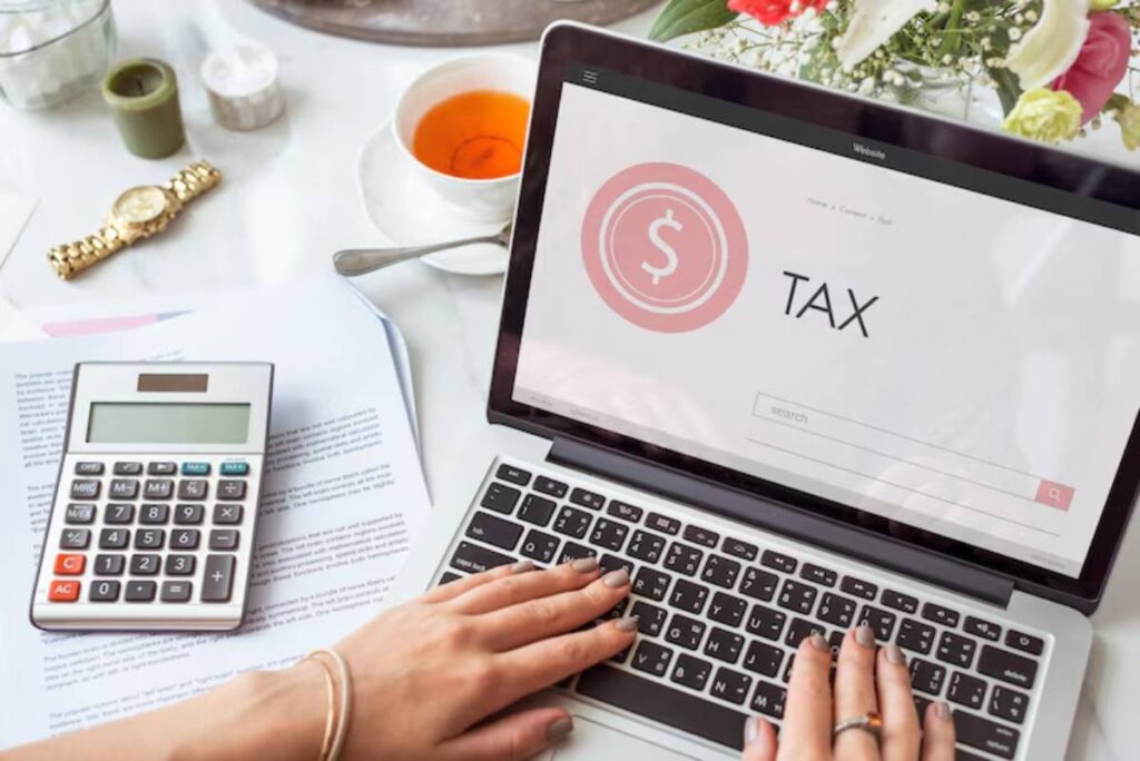 A hand typing on a laptop displaying a tax search page, with a calculator, papers, tea, and flowers on a table.