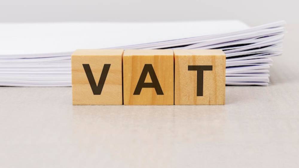 Wooden blocks spelling 'VAT' are placed in front of a stack of white papers on a light surface. The scene conveys a professional and financial theme.