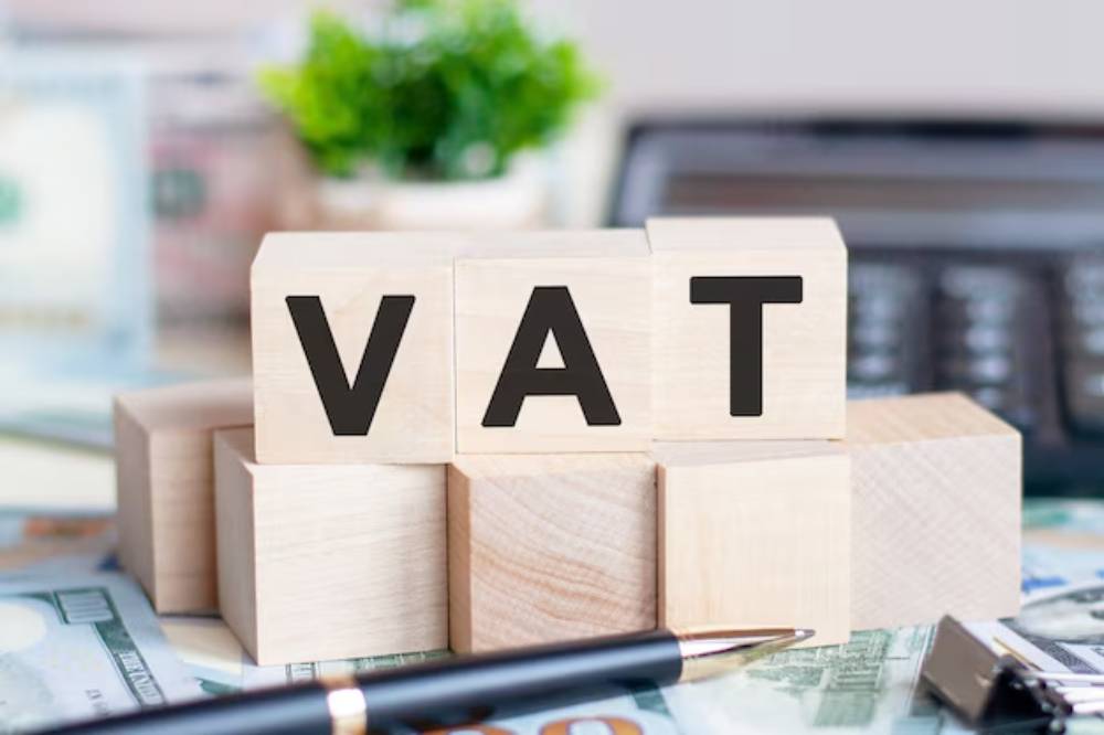 Wooden blocks spelling 'VAT' on a desk with cash and a pen, symbolizing tax concepts in a financial setting.
