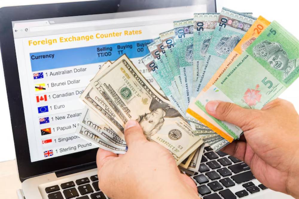 Hands holding various banknotes in front of a laptop displaying foreign exchange counter rates for different currencies.