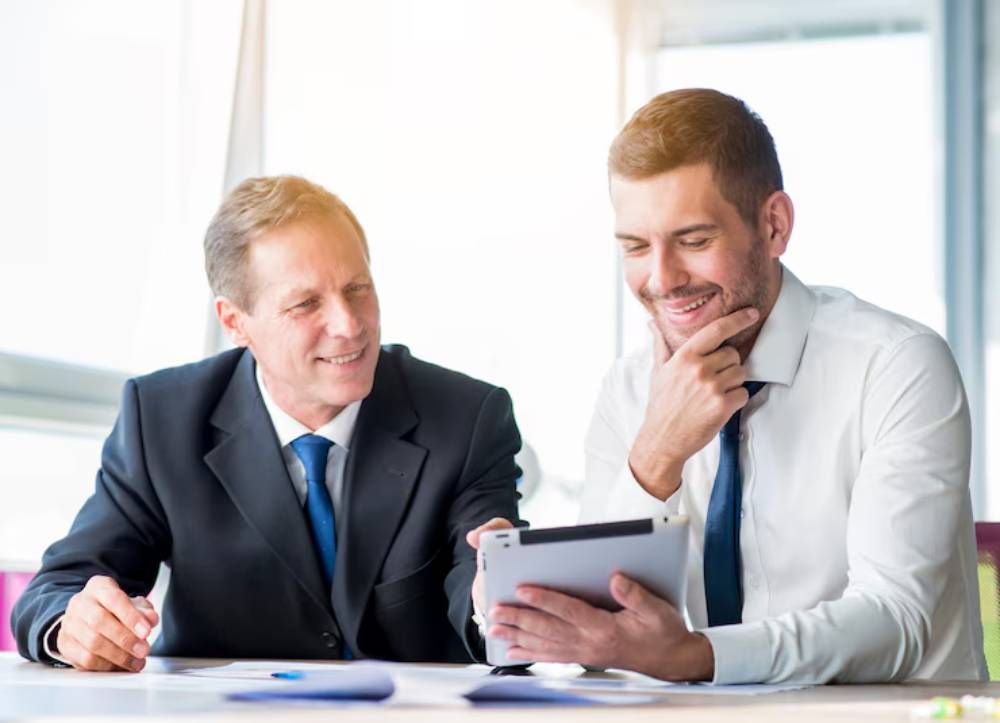 Two businessmen in formal attire are engaged in discussion, reviewing documents on a tablet at a bright, modern office.