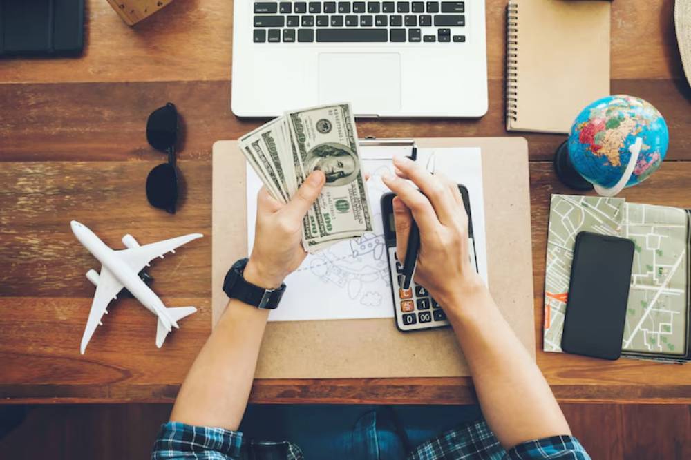 A person counts cash  surrounded by a laptop, airline model, map, globe, and calculator on a wooden desk.