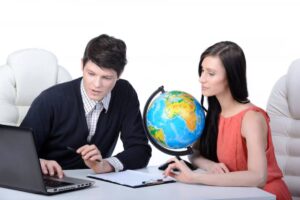 A man and a woman sit at a desk with a laptop and a globe, discussing plans or ideas in a bright, modern office setting.