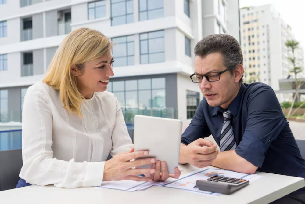 Two professionals review documents and a tablet at an outdoor table, with modern buildings and greenery in the background.