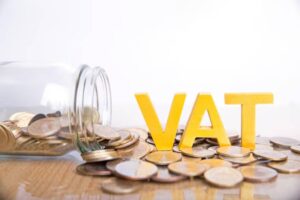 A glass jar spilling coins beside bold yellow letters spelling 'VAT,' symbolizing value-added tax and savings.