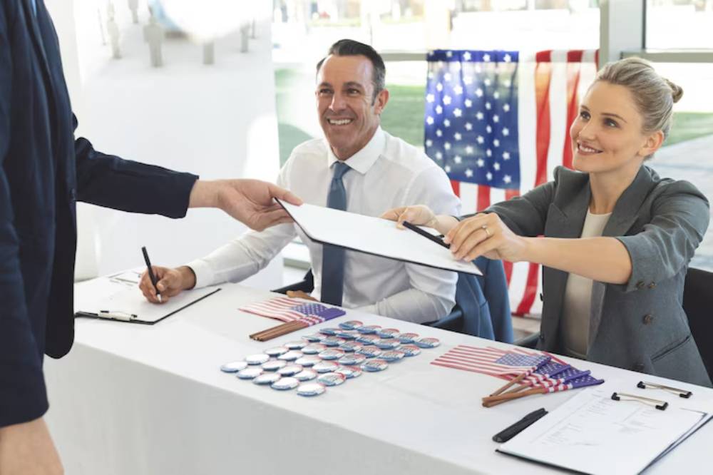A person hands a clipboard to two officials at a table, surrounded by American flags and election-themed buttons and pens.