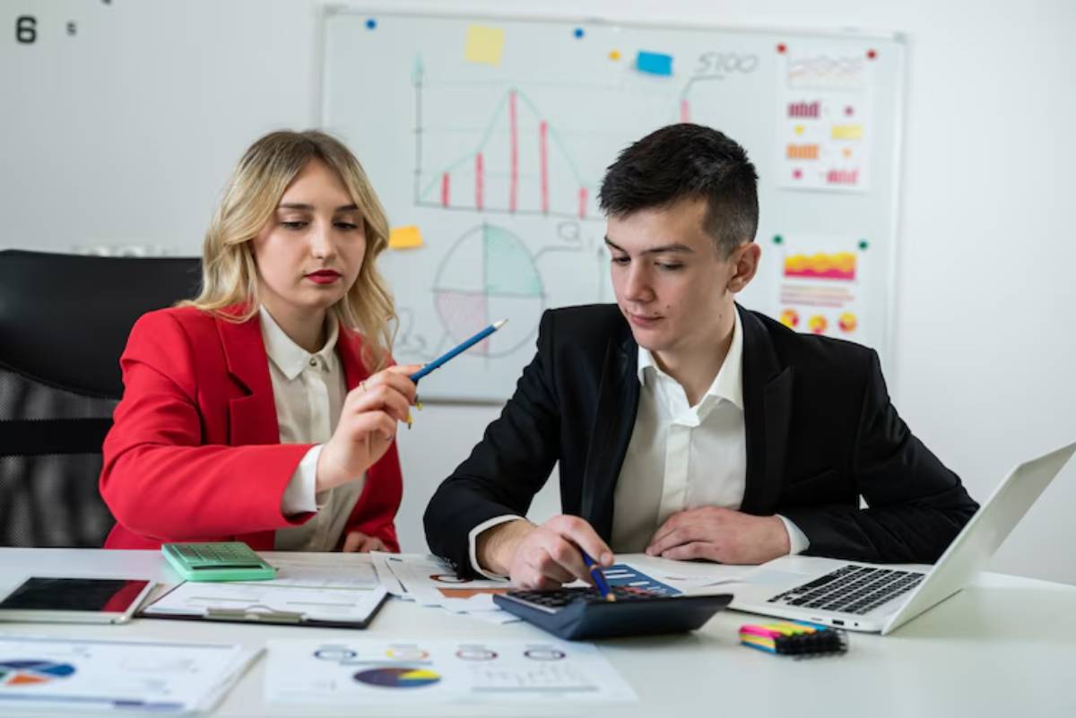 A woman in a red blazer points at documents while a man in a black suit examines a calculator and papers on a desk with a laptop.