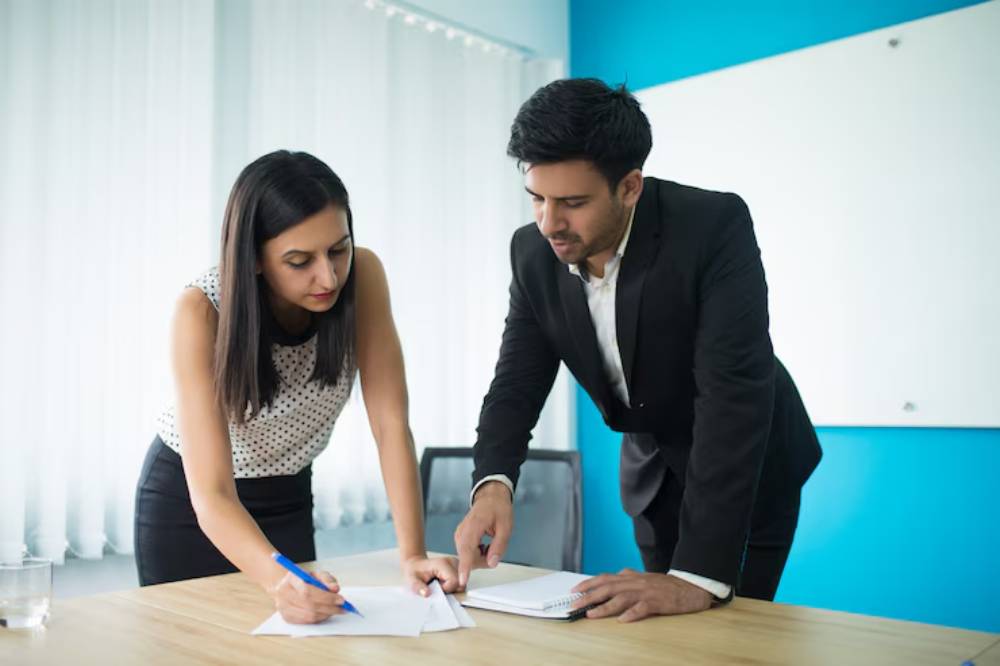 A woman in a polka dot blouse writes on paper while a man in a suit points at notes on a notepad during a business meeting.