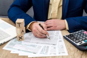 A person in a blue blazer is filling out tax forms with a pen, while blocks spelling 'TAX' and a calculator are nearby.