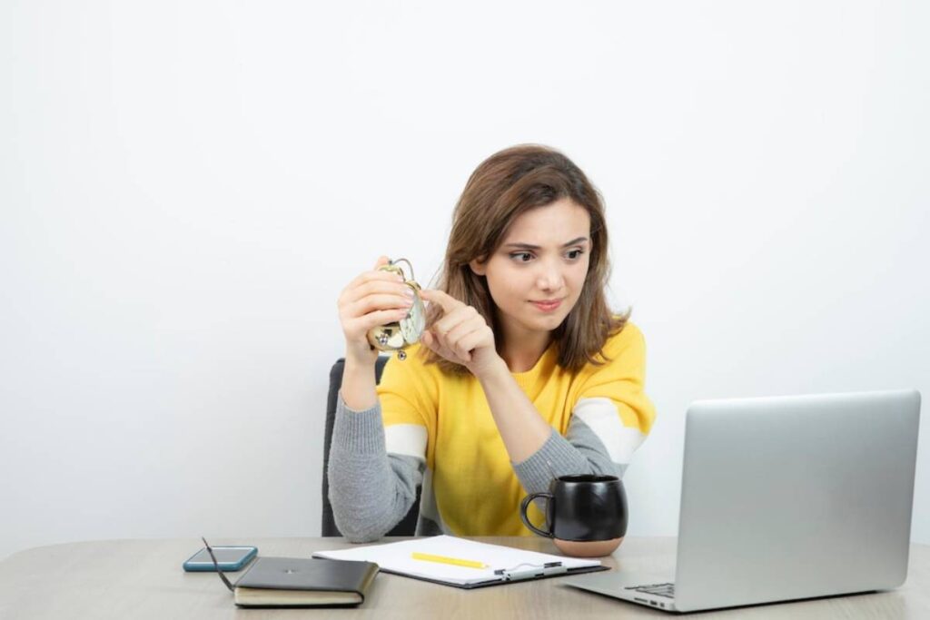A person in a yellow sweater sits at a desk with a laptop, holding an alarm clock and surrounded by a notebook, phone, and coffee cup.