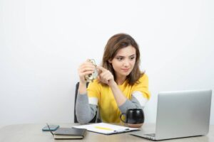 A person in a yellow sweater sits at a desk with a laptop, holding an alarm clock and surrounded by a notebook, phone, and coffee cup.