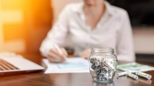 A glass jar filled with rolled dollar bills sits on a table next to a laptop, as someone writes on paper.