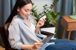 A person sits in a chair holding a tablet and a credit card, surrounded by greenery and soft, natural lighting.