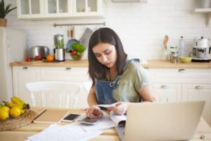 A person sits at a kitchen table, reviewing documents and using a calculator, surrounded by fruits and a laptop.