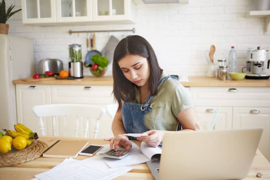 A person sits at a kitchen table, reviewing documents and using a calculator, surrounded by fruits and a laptop.