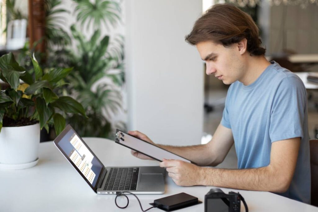 A person in a blue shirt sits at a desk, using a laptop and holding a clipboard, with a camera and a plant in the background.