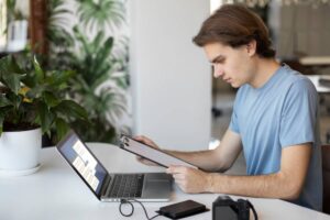 A person in a blue shirt sits at a desk, using a laptop and holding a clipboard, with a camera and a plant in the background.