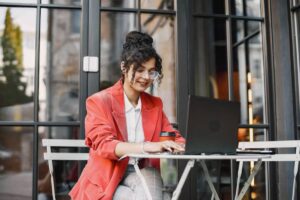 A person wearing a red blazer sits at a table outdoors, focused on their laptop, with a coffee cup beside them.