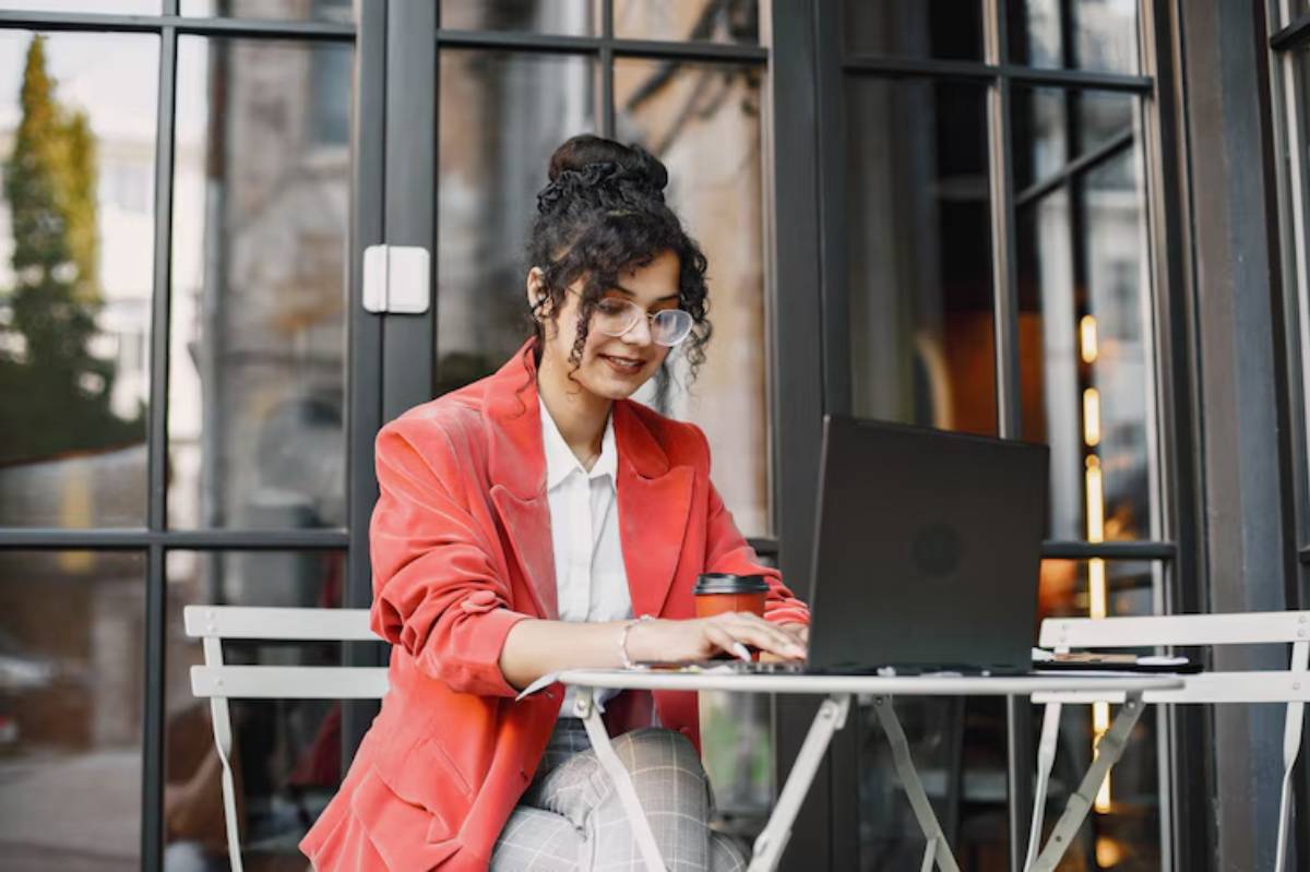 A person wearing a red blazer sits at a table outdoors, focused on their laptop, with a coffee cup beside them.