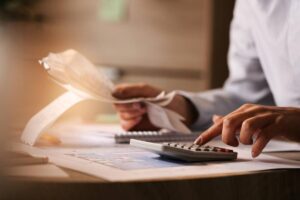 A person calculating expenses with a calculator, holding receipts, and reviewing financial documents on a desk.