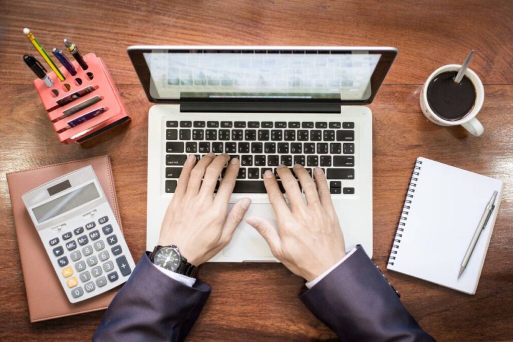A person's hands typing on a laptop keyboard, surrounded by a calculator, notebook, pens, and a coffee cup on a wooden desk.