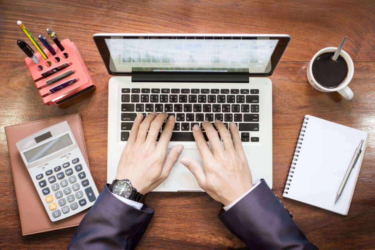 A person's hands typing on a laptop keyboard, surrounded by a calculator, notebook, pens, and a coffee cup on a wooden desk.