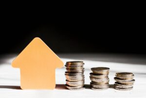 A paper house icon stands beside stacks of coins in varying heights, symbolizing financial growth.