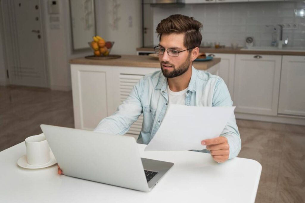 A person sitting at a table in a modern kitchen, focusing on a laptop while holding documents, with a coffee cup nearby.
