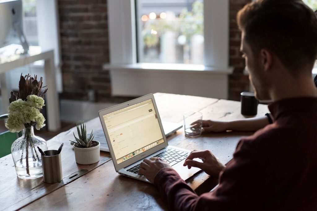 A person typing on a laptop at a rustic wooden table, surrounded by potted plants and a glass of water in a cozy environment.