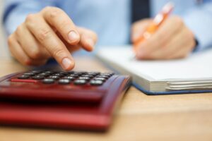 A hand pressing buttons on a red calculator, with another hand holding a pen writing on a blank notebook page.