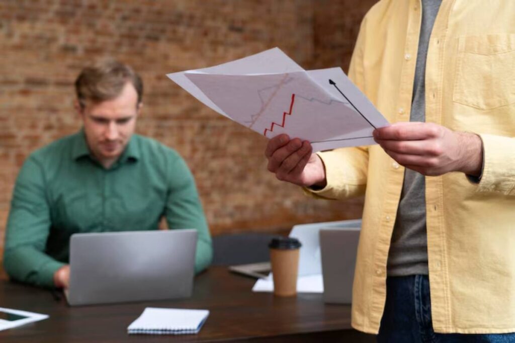 A person holding documents with graphs stands in an office meeting, while another individual works on a laptop at the table.