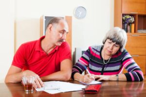 A woman in a striped sweater writes notes while a man in a red shirt assists at a table with documents and drinks.