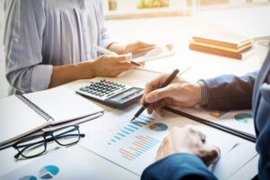 Two professionals analyze charts and data on a table, using a calculator and notebooks, in a bright office setting.