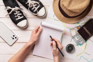 Aerial view of a hand holding a pencil over a notepad, surrounded by a phone, sneakers, a map, a compass, cash, and a straw hat.