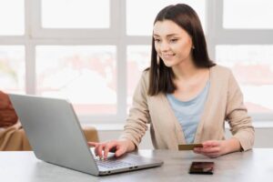 A woman sits at a desk using a laptop, holding a credit card in her right hand and a smartphone nearby. Natural light shines through a window.