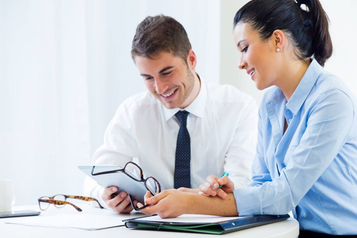 Two professionals collaborate at a table, reviewing a tablet and notes, with glasses and a pen nearby. Soft, light-filled background.