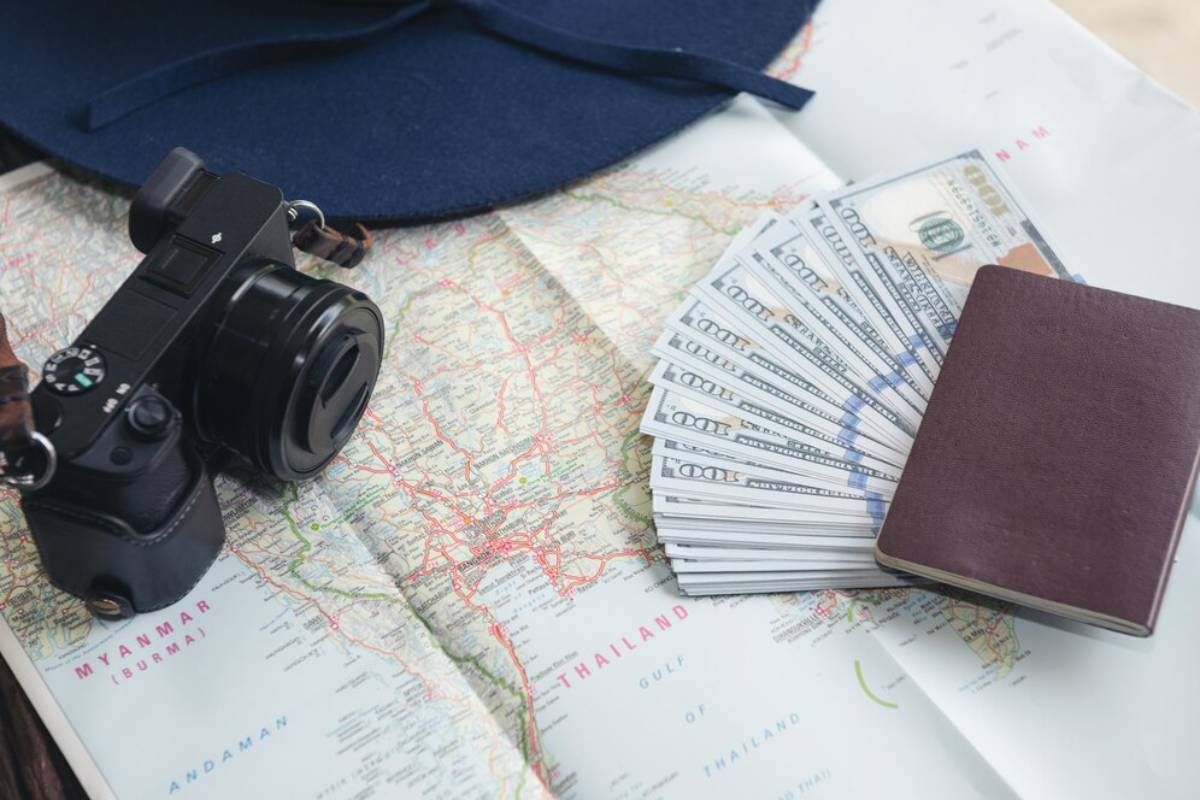 A black camera, folded map, stack of money, a passport, and a blue hat arranged on a travel-themed table, ready for adventure.