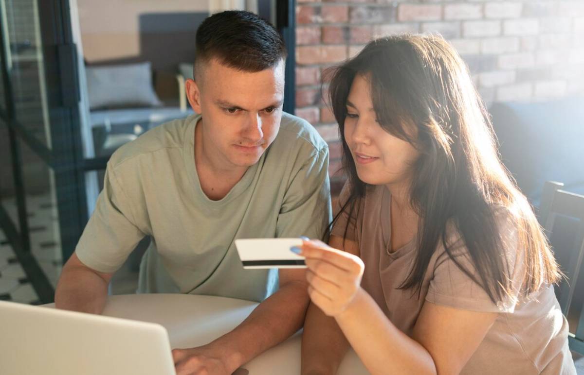 A young couple sits at a table, focused on a laptop, while one of them holds a credit card.