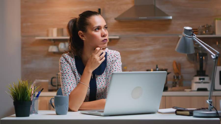 A woman sits at a desk in a cozy kitchen, deep in thought, with a laptop, a mug, and potted plants surrounding her.