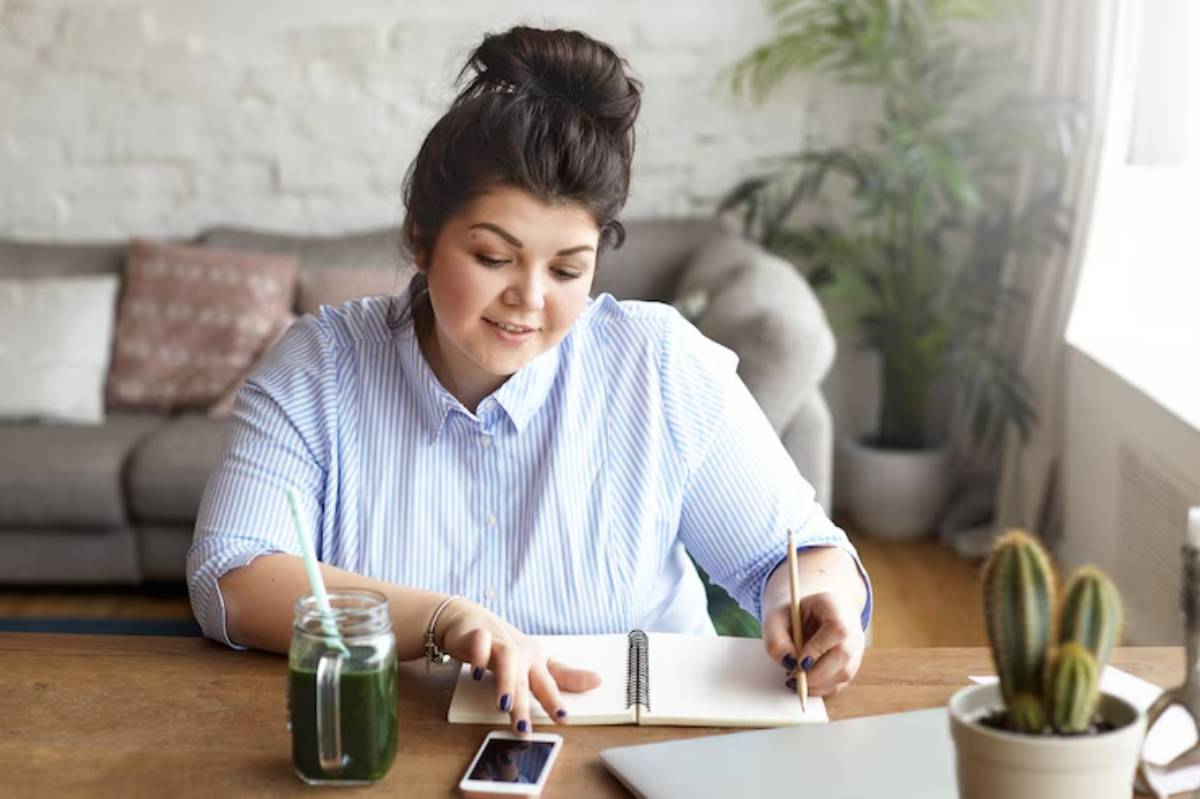 A person sits at a wooden desk with a notebook, pen, and drinks, surrounded by plants and a cozy living room setting.