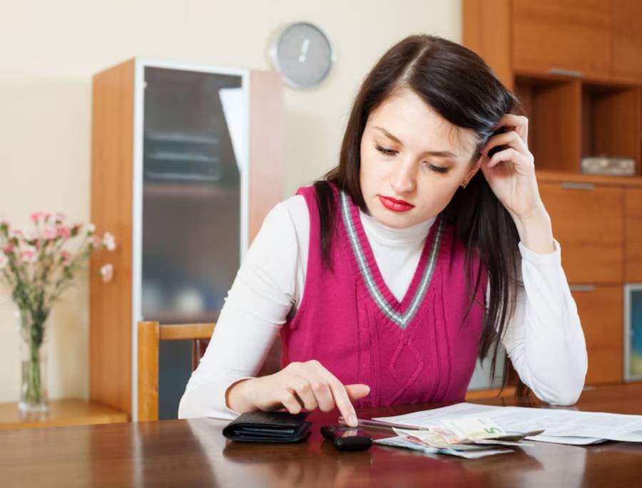 A woman in a pink sweater sits at a table with a cellphone, wallet, bills, and cash, looking concerned at her finances.