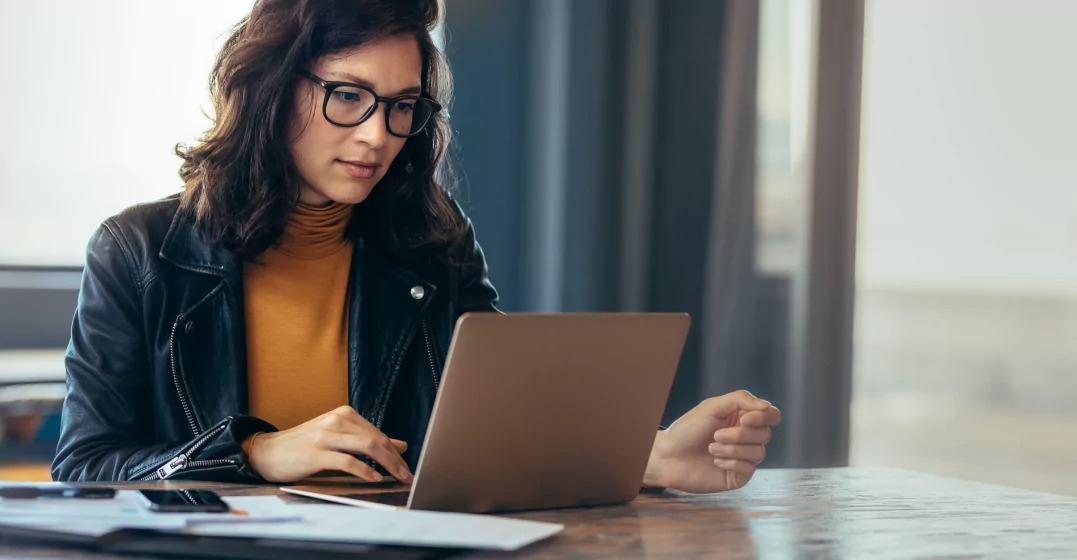 A person in a leather jacket sits at a wooden table, working on a laptop with stationery and a smartphone nearby.