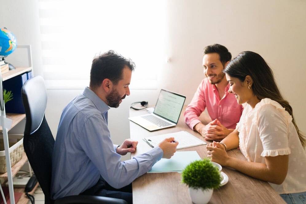 A professional meeting occurring between three individuals at a desk with a laptop, documents, and a small potted plant.