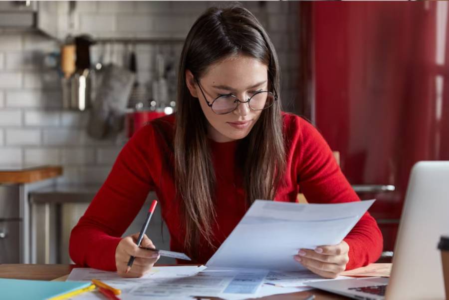 A person in a red sweater sits at a wooden desk, examining documents and writing notes while using a laptop in a cozy kitchen setting.