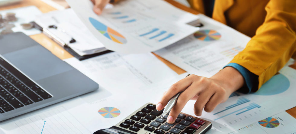 A person in a yellow jacket calculates finances with a calculator, surrounded by documents and charts on a wooden desk.