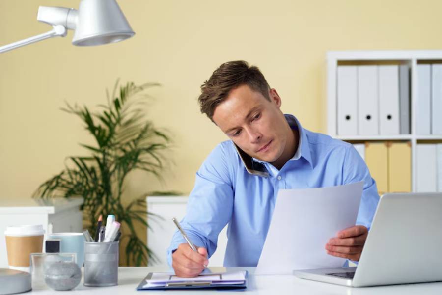 A person in a light blue shirt reviews documents at a desk with a laptop, plants, and office supplies in a bright workspace.