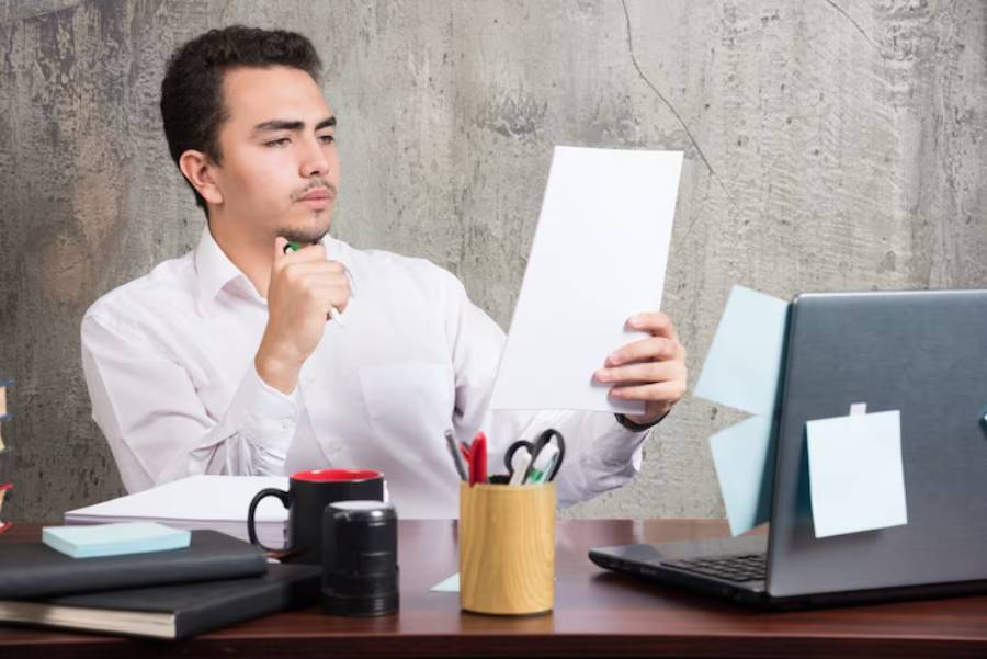 A person sitting at a desk, holding a sheet of paper, with a laptop, notepad, and various office supplies in a modern workspace.