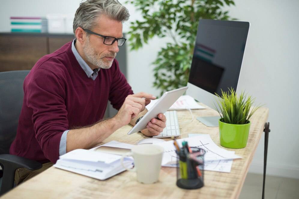 A man in a burgundy sweater uses a tablet at a wooden desk cluttered with papers, a cup, and a small green plant.