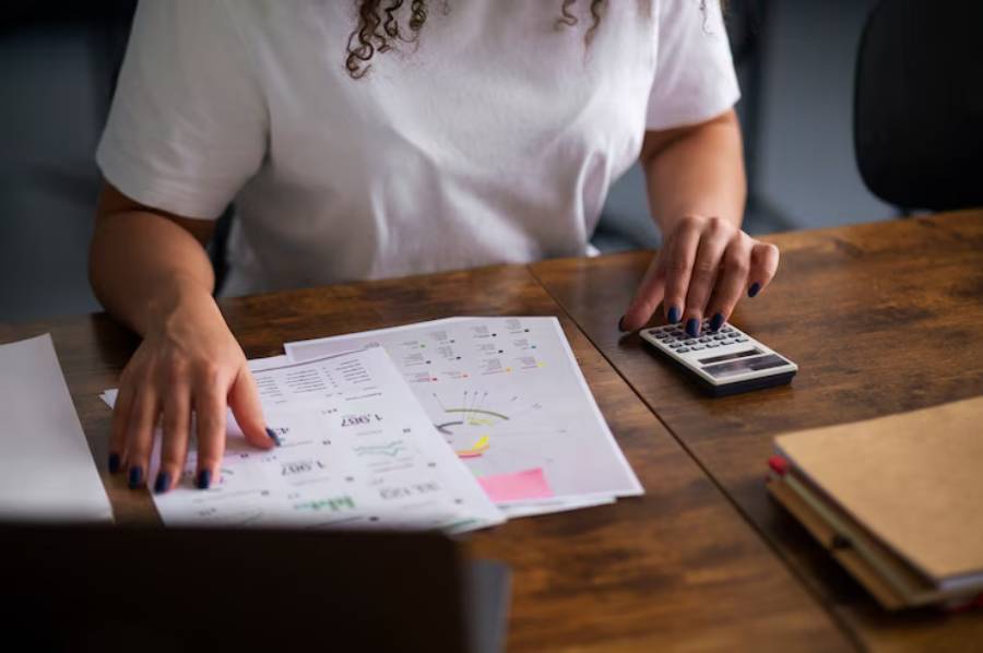 A person reviews financial documents and charts while using a calculator on a wooden table, focused on their work.