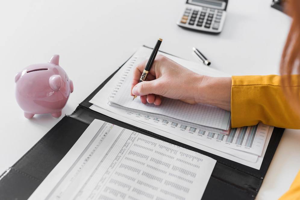A person in a yellow shirt writes on a notepad next to a pink piggy bank and financial documents, with a calculator in the background.